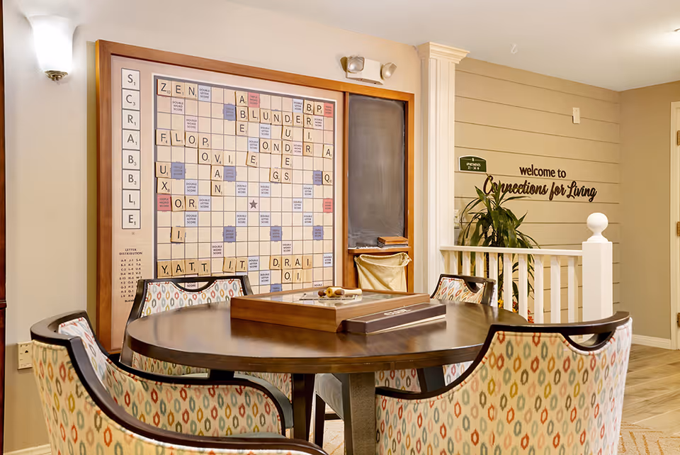 A cozy game room with a round wooden table surrounded by four patterned chairs. On the wall is a large Scrabble board with tiles arranged to form words. Next to the board is a chalkboard and a small basket. The wall has a sign that reads 'welcome to Connections for Living' with a green room number sign above it. The room has warm lighting and a welcoming atmosphere.