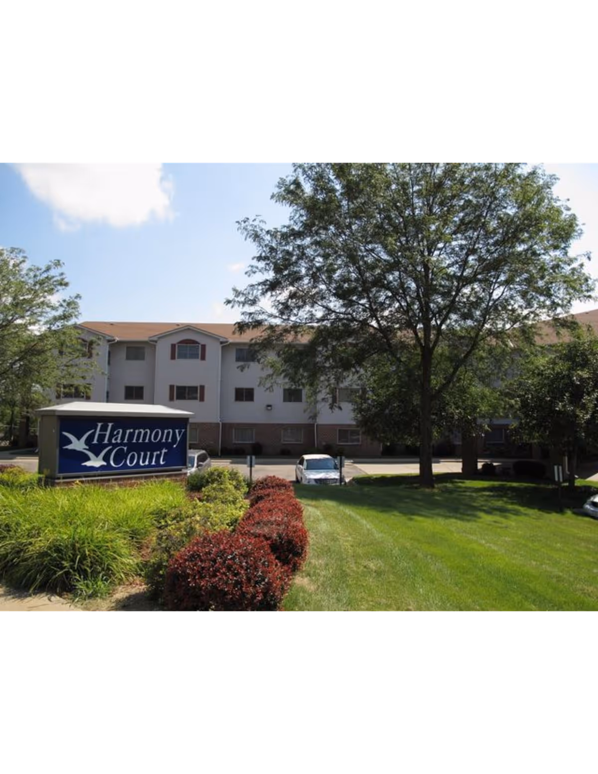 Exterior view of Harmony Court senior living facility in Council Bluffs, IA, showing a three-story building with a beige and brick facade, surrounded by green grass, trees, and landscaped bushes. A sign in front reads 'Harmony Court' with a bird logo.