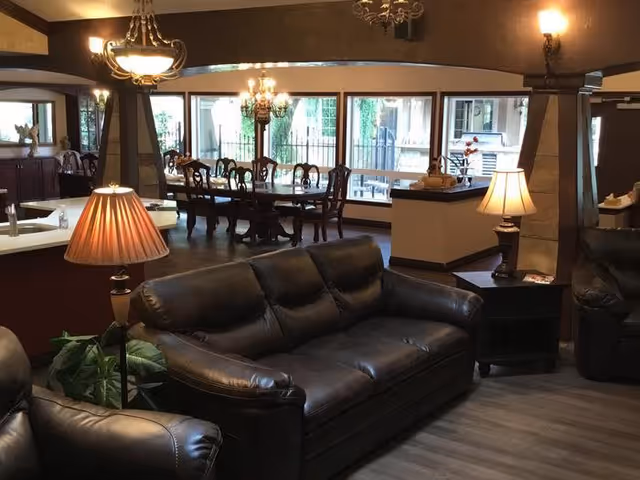 Interior view of a senior living facility common area featuring dark leather sofas, a wooden dining table with chairs, multiple lamps providing warm lighting, large windows letting in natural light, and decorative columns.