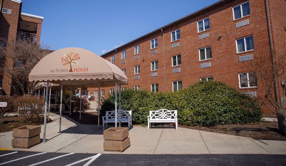 Exterior view of Autumn House West, showing a brick building with multiple windows, a covered entrance canopy with the Autumn House logo, white benches, and some greenery under a clear blue sky.