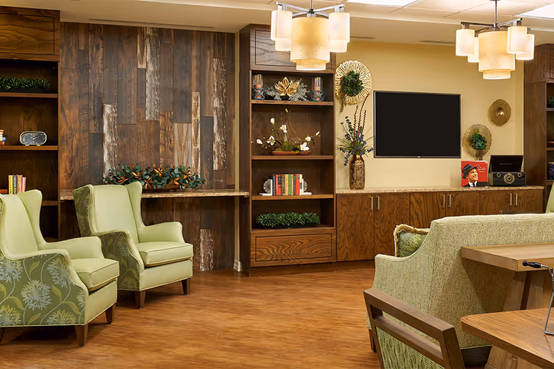 A cozy senior living common area with green upholstered armchairs and a sofa, wooden bookshelves filled with books and decorative plants, a flat-screen TV mounted on a beige wall, and warm pendant lighting overhead.