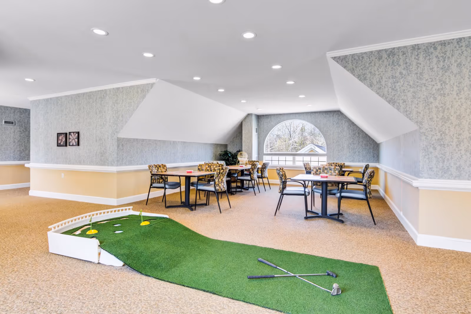 A spacious indoor room with beige carpet and light blue patterned wallpaper, featuring several tables and chairs arranged for seating. In the foreground, there is a small indoor putting green with two golf clubs resting on it. The room has a large arched window letting in natural light.