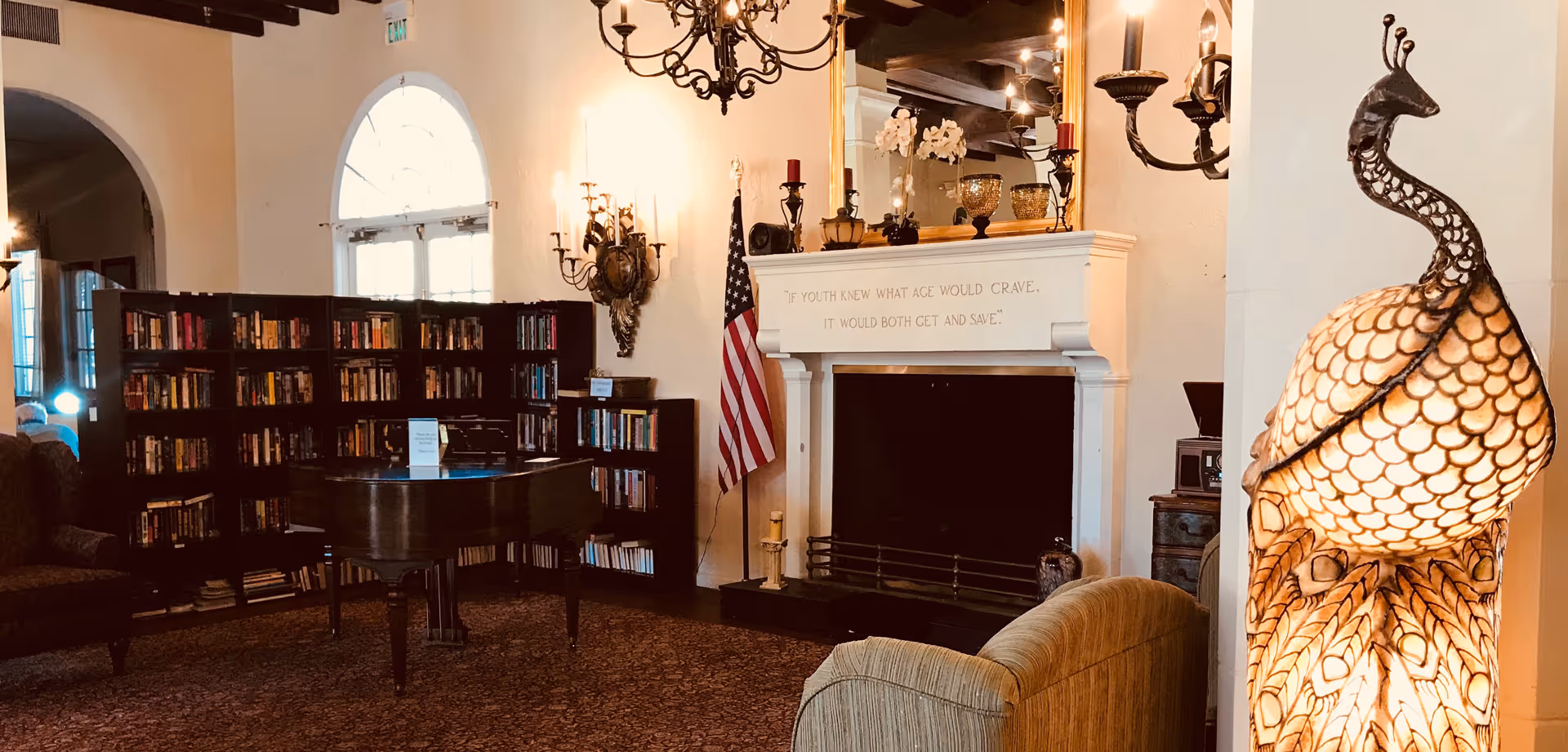 A cozy lounge with bookshelves and a grand piano facing a fireplace under a chandelier.