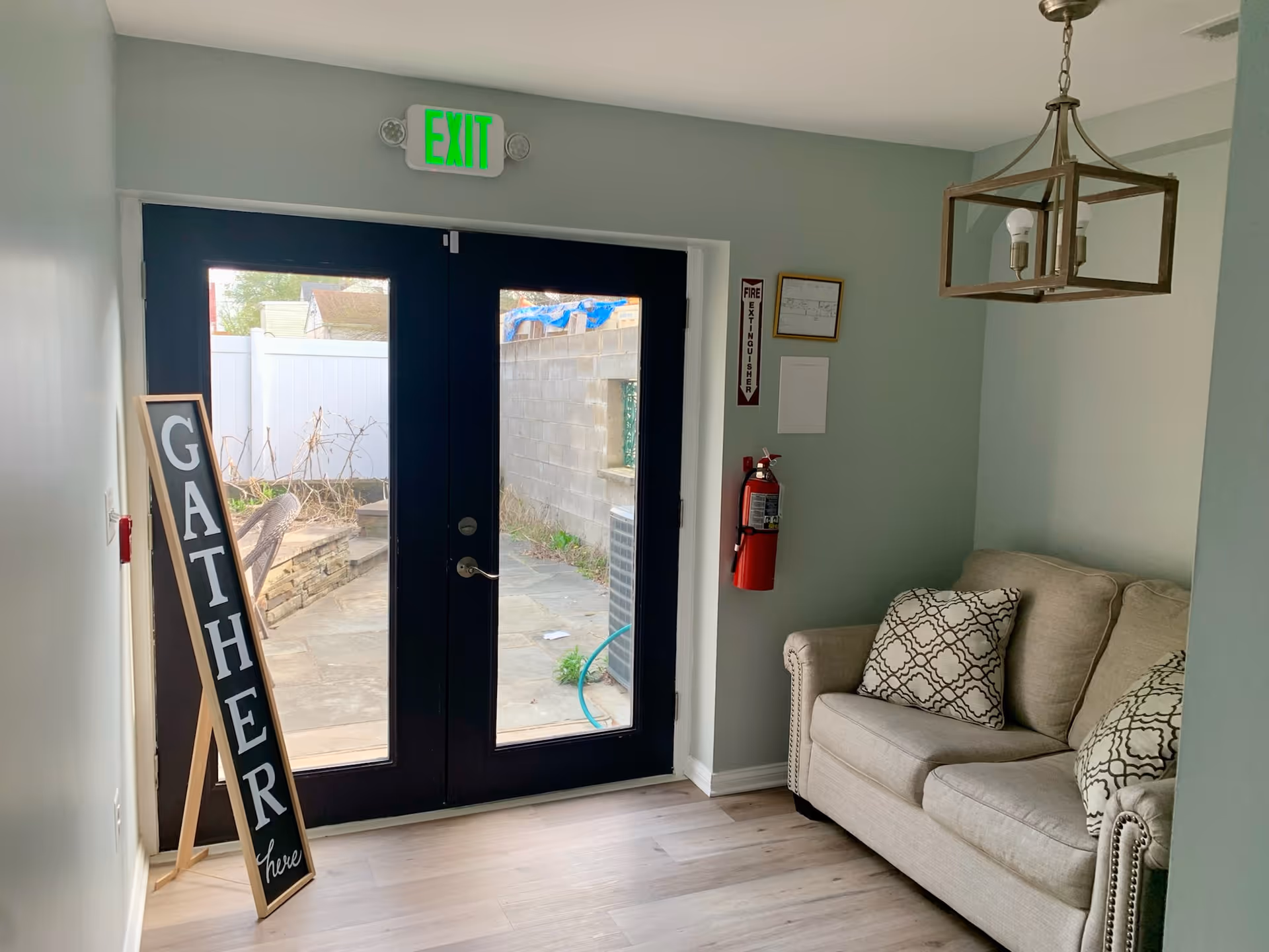 Small entry seating area with a beige loveseat and patterned pillows, a pendant light, an 'EXIT' sign above double glass doors, and a freestanding 'GATHER' sign.
