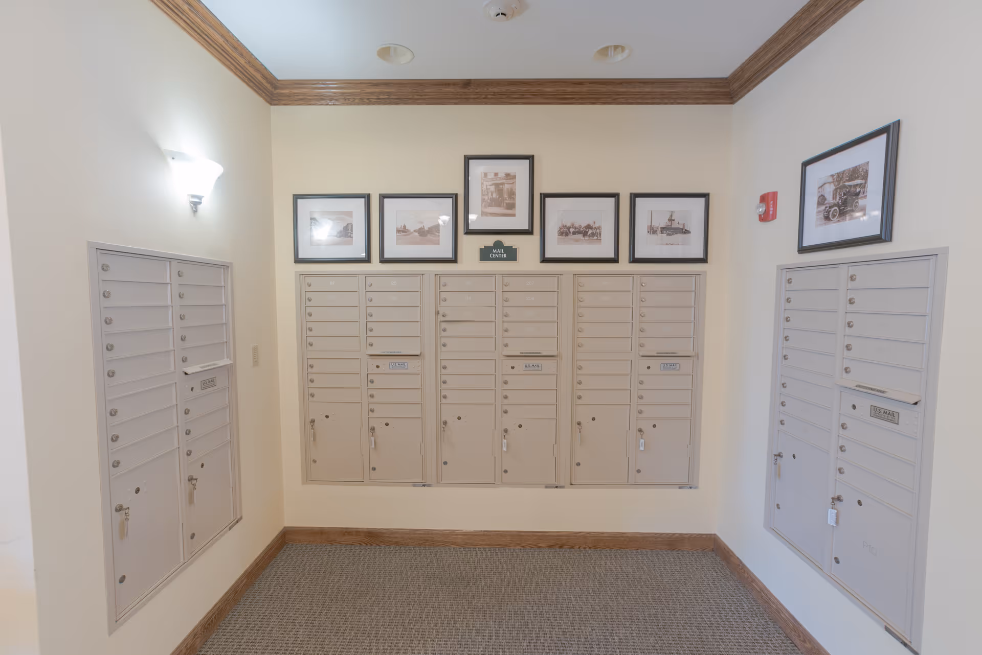 Interior view of a mail center room with multiple beige mailboxes mounted on three walls. Above the mailboxes are framed black and white photographs. The room has beige walls, a carpeted floor, and wooden trim along the ceiling and floor edges. A wall-mounted light fixture is visible on the left wall.