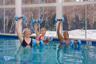 Three senior women in swimsuits participating in a water exercise class in an indoor swimming pool, each holding blue water dumbbells and raising one arm above their heads. Large windows in the background show a snowy outdoor scene with trees.