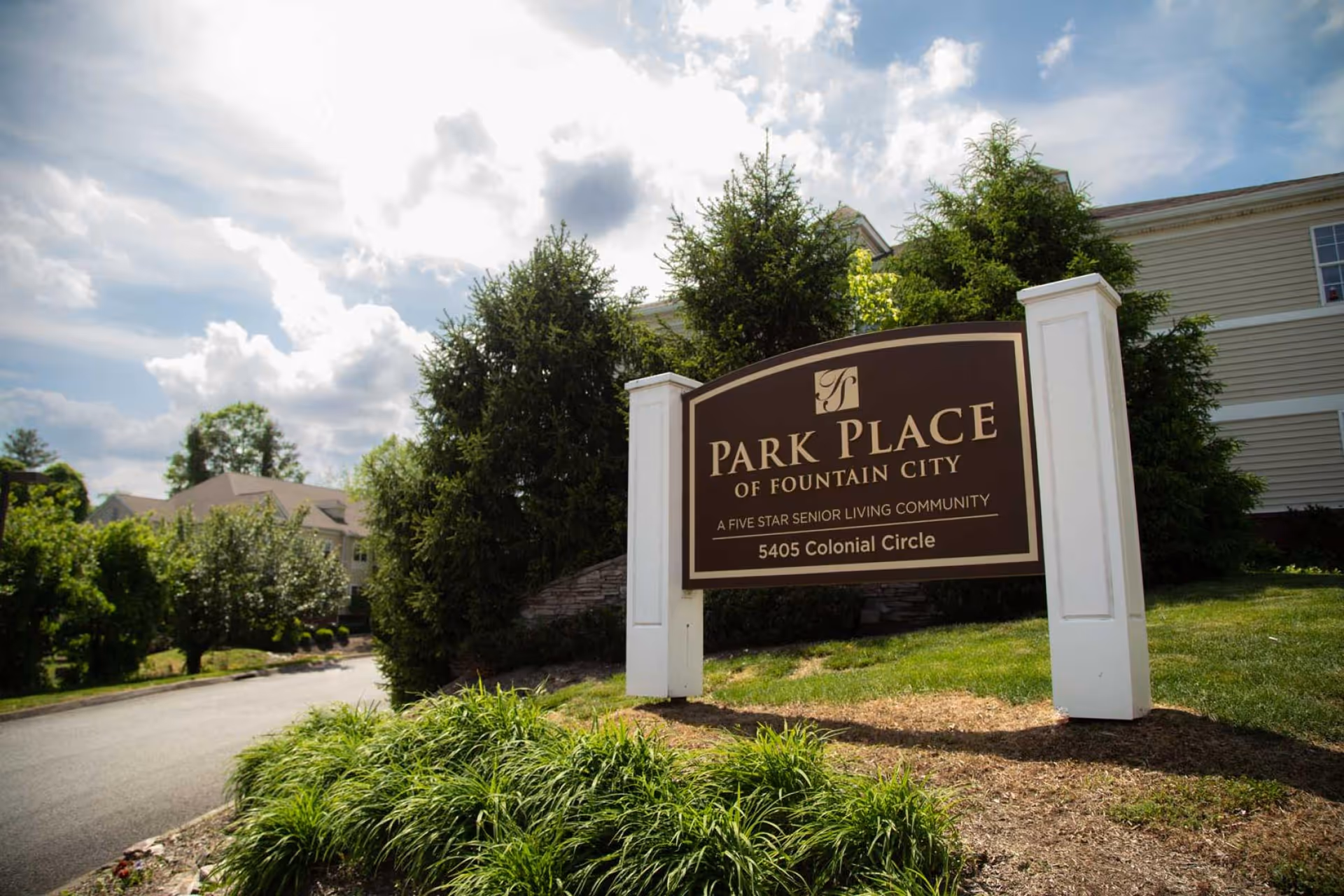 Outdoor view of the entrance sign for Park Place of Fountain City, a senior living community, surrounded by greenery and trees with a partly cloudy sky in the background.