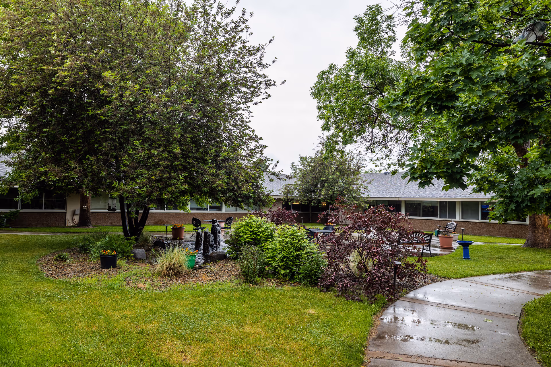 A lush outdoor garden area at Yellowstone River Nursing and Rehabilitation featuring green grass, various trees and shrubs, a small water fountain, potted plants, and outdoor seating with chairs and tables. A wet concrete pathway curves through the garden, and a single-story building with large windows is visible in the background.