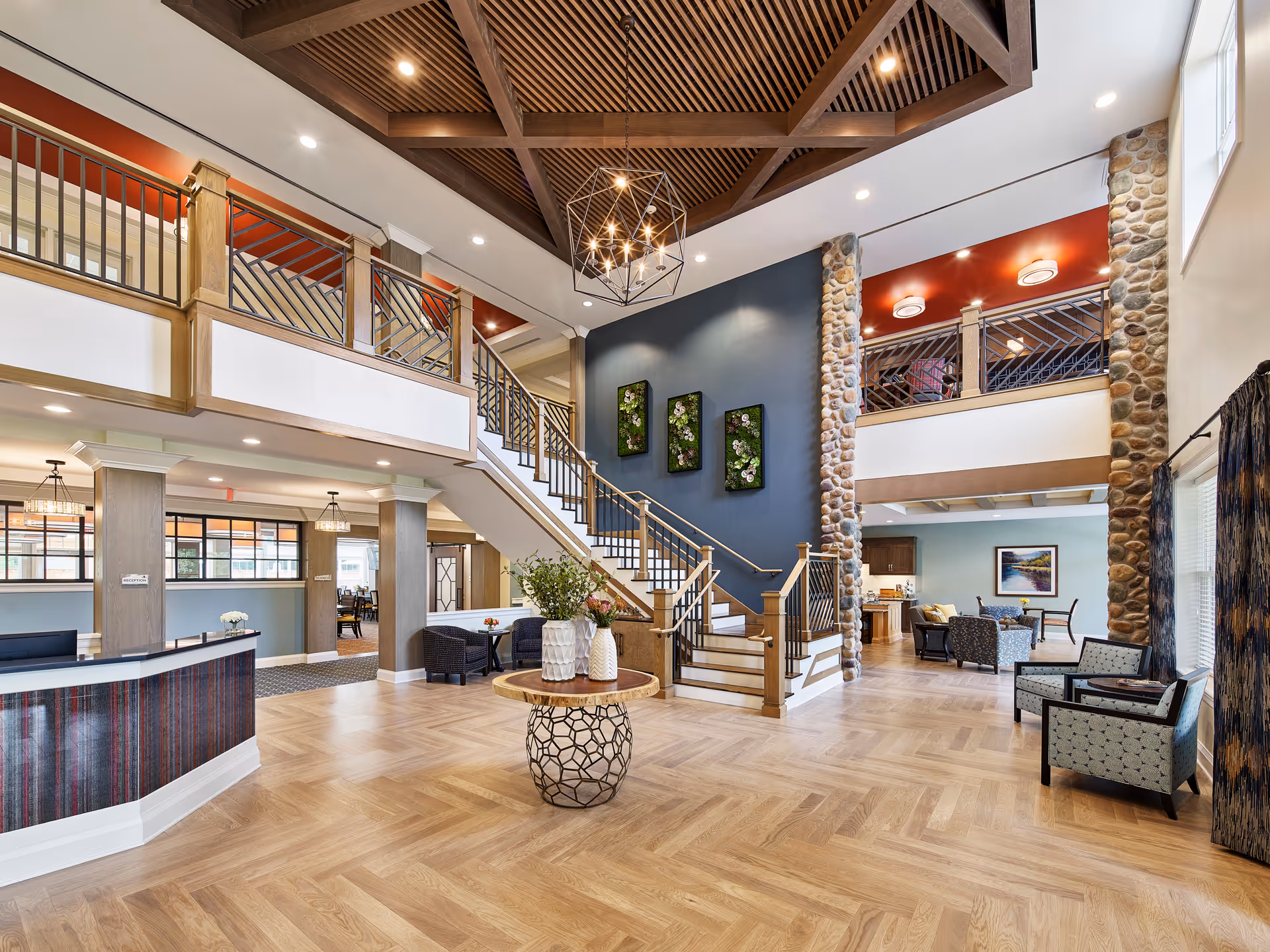 Spacious two-story senior living facility lobby with a central staircase, reception desk, seating areas, and decorative ceiling.
