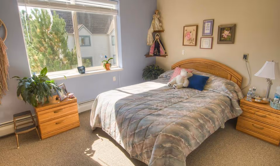 A bright bedroom in an assisted living facility with a large window letting in natural light. The room features a double bed with a patterned comforter, a stuffed animal, and pillows. There are two wooden nightstands on either side of the bed, one with a lamp and water bottles, and the other with a plant and framed photo. The walls have framed pictures and religious figurines on corner shelves. A small step stool is next to one nightstand.
