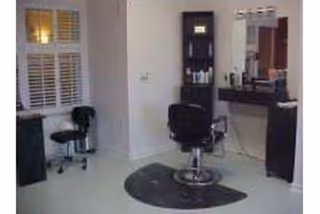 Interior view of a salon area with a black salon chair on a protective mat in front of a mirror and counter with hair care products. There is a small stool and a window with white shutters on the left side.