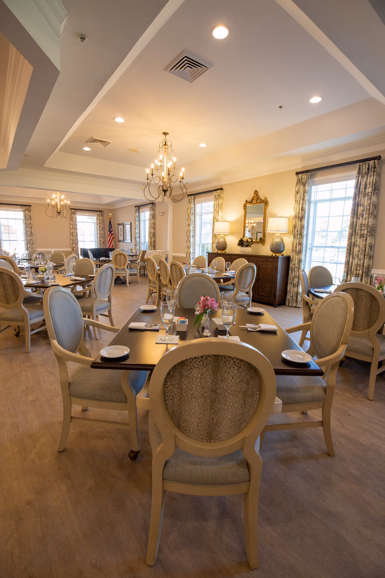 A well-lit dining room with multiple round and rectangular tables set with plates, glasses, napkins, and silverware. The room features beige upholstered chairs, large windows with patterned curtains, chandeliers hanging from the ceiling, and a wooden sideboard with lamps and a mirror above it. An American flag is visible in the background near the windows.