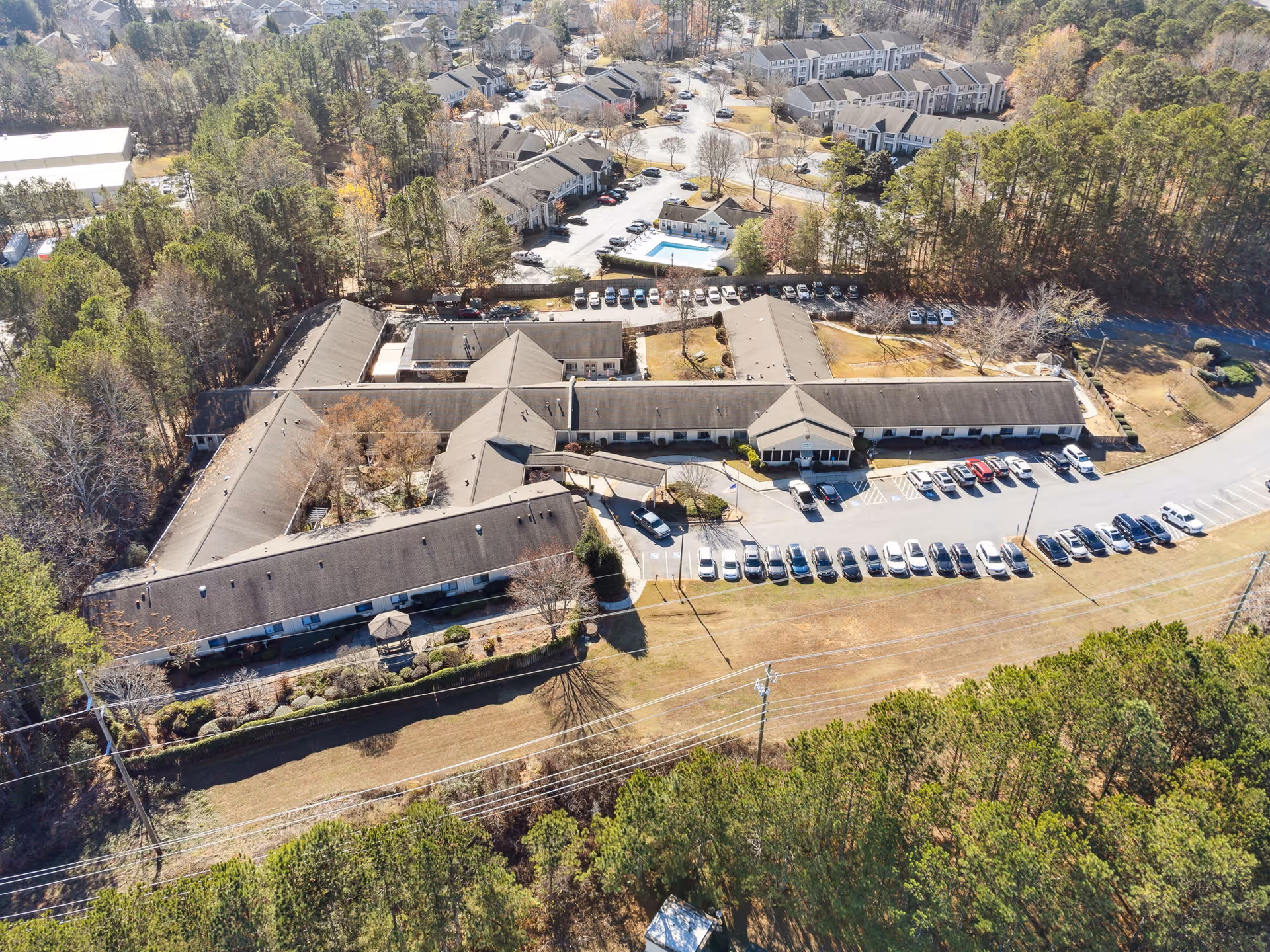 Aerial view of Cumming Health & Rehab facility showing a large building complex with multiple wings surrounded by trees and parking lots filled with cars. Residential houses and a swimming pool are visible in the background.