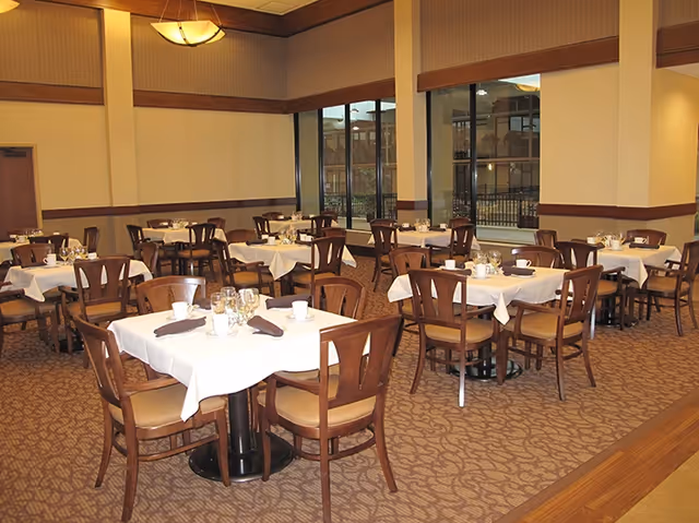 A dining room with multiple tables covered in white tablecloths, each set with glasses, cups, and napkins. Wooden chairs with cushioned seats surround the tables. Large windows on one side show an outdoor view, and the room has warm lighting and carpeted floors.