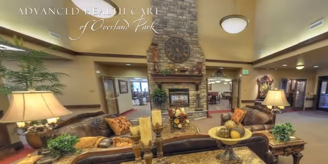 A cozy living room area in a senior living facility with leather sofas, decorative pillows, a stone fireplace with a large clock above it, and various decorative items including candles and plants. The room has warm lighting and an inviting atmosphere.