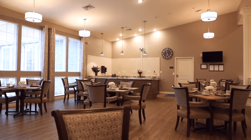 A well-lit dining room with multiple round tables and cushioned chairs arranged neatly. Each table is set with cups, plates, and floral centerpieces. Large windows with curtains allow natural light to fill the room. The back wall features a counter with decorative items, a clock, and a mounted television.