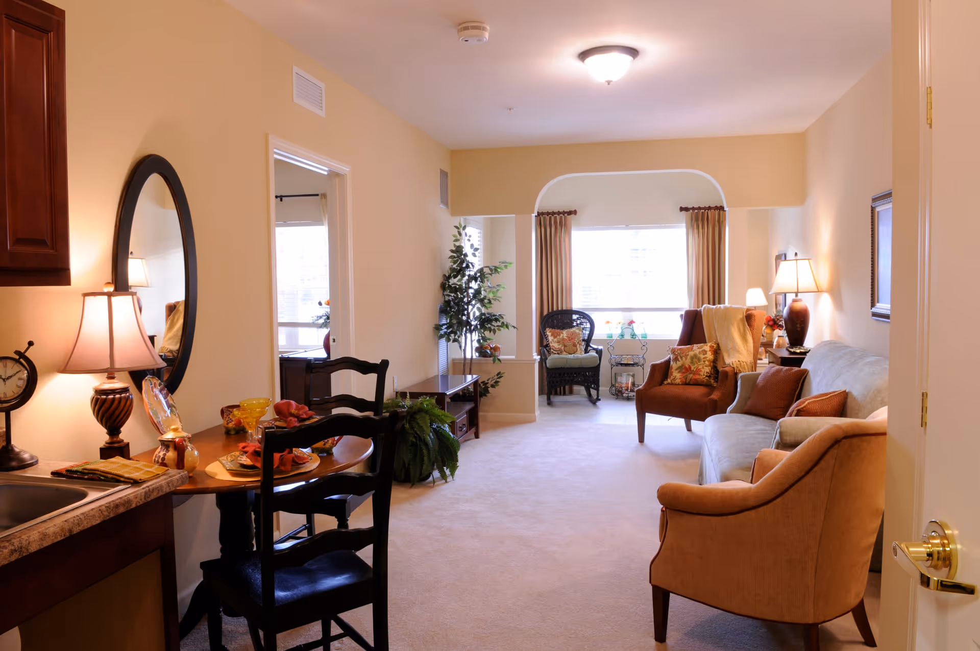 A cozy living room and dining area in a senior living facility with beige walls and carpet. The living room features a sofa, armchairs, a side table with a lamp, and a large window with curtains. The dining area has a small round table set for two with chairs, a lamp, a clock, and a round mirror on the wall. There is also a potted plant near the window.