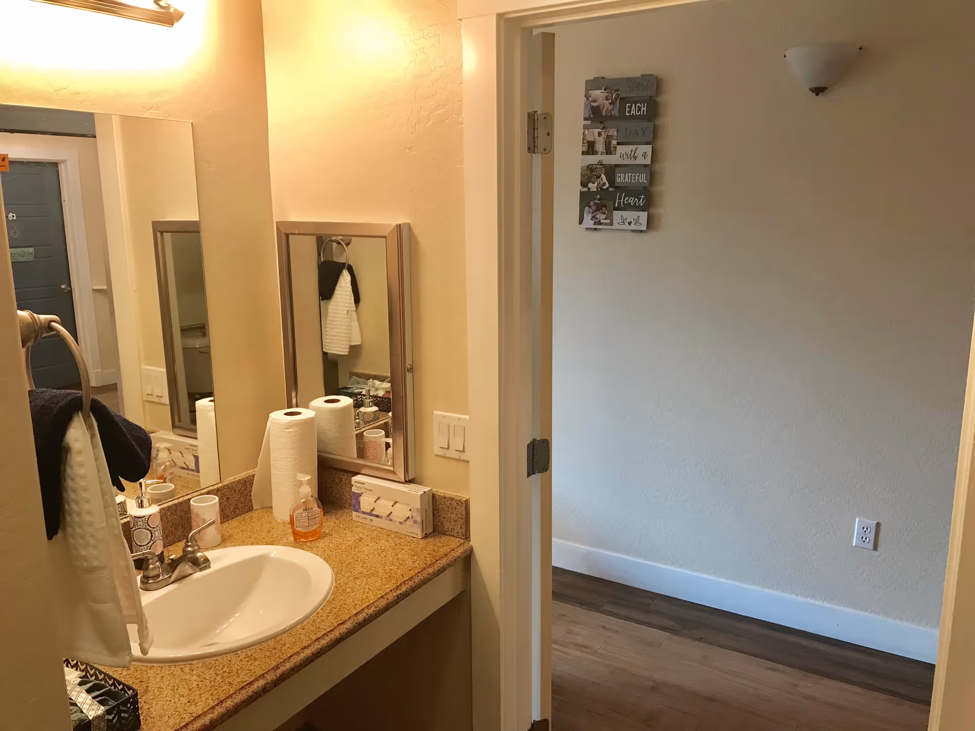 Bathroom vanity with sink, mirror, hand soap and paper towels beside an open doorway into an empty room.
