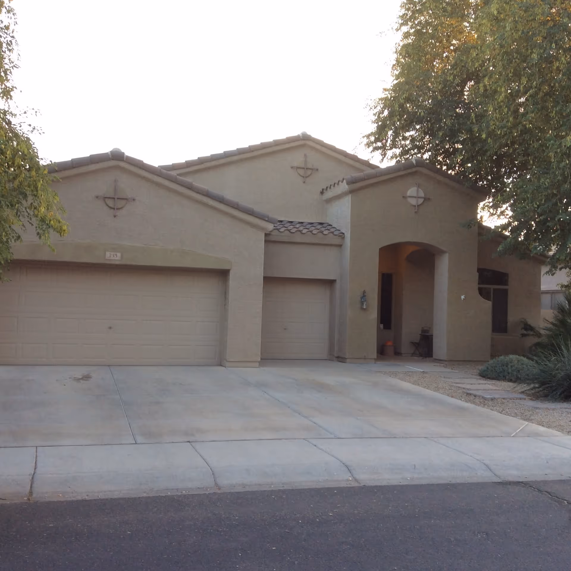 Exterior view of a beige stucco house with a three-car garage, a tiled roof, and a small covered porch area. There are trees and shrubs around the house, and a concrete driveway leading to the garage doors.
