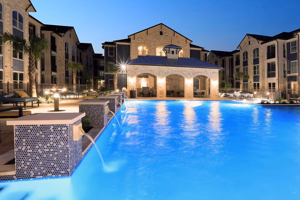 Illuminated outdoor swimming pool at night surrounded by multi-story residential buildings and poolside seating.