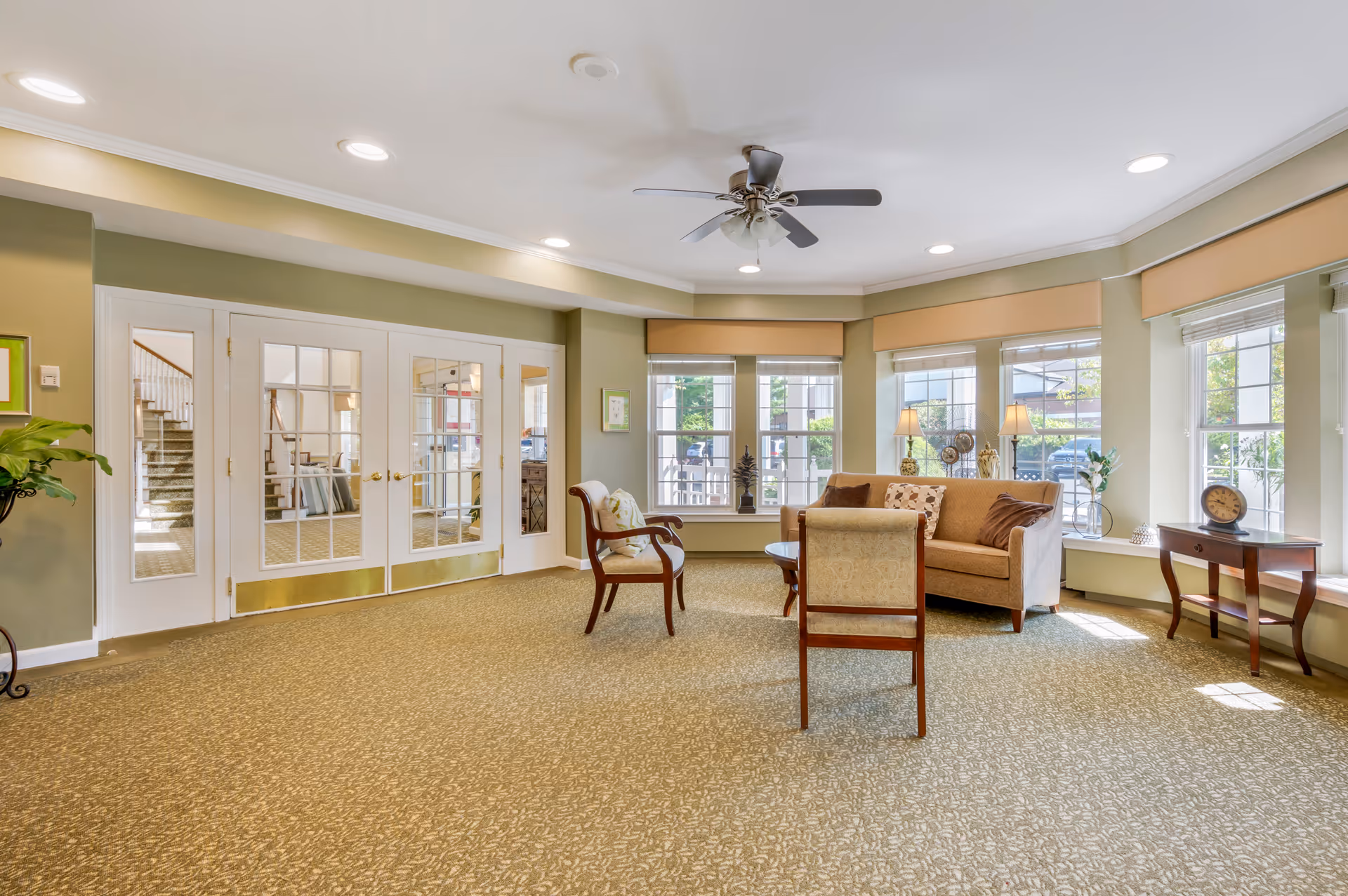 A bright and spacious living room area with beige carpet, light green walls, and large windows letting in natural light. The room features a beige sofa with decorative pillows, two wooden chairs with cushions, a round wooden coffee table, and a small side table with a clock. There is a ceiling fan with lights and recessed lighting in the ceiling. Double glass doors lead to another room with a staircase visible.