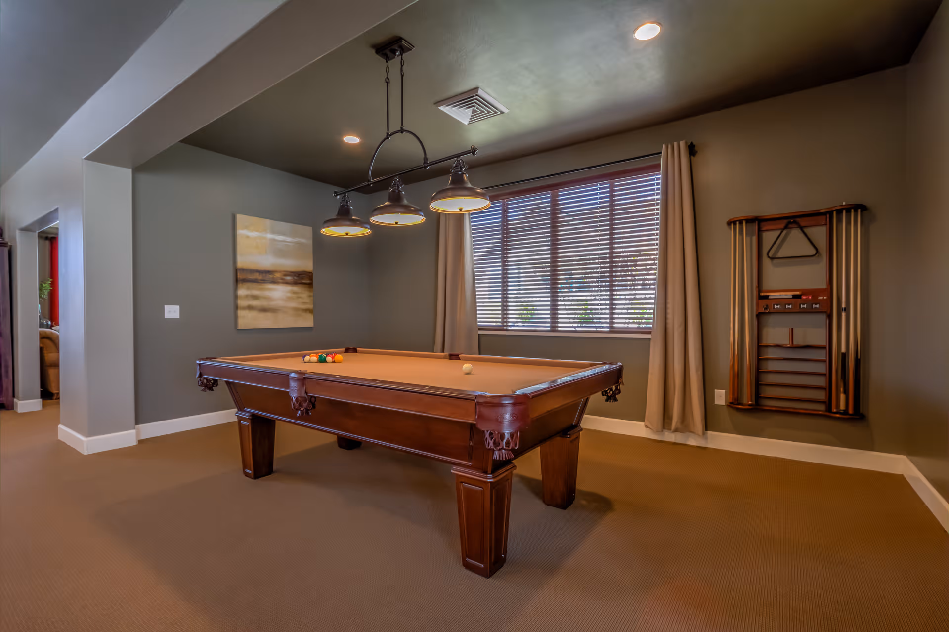 A well-lit game room with a brown pool table in the center, a set of billiard balls on the table, a hanging light fixture above, a window with blinds and beige curtains, a wall-mounted rack holding pool cues and a triangle rack, and a piece of abstract art on the wall.