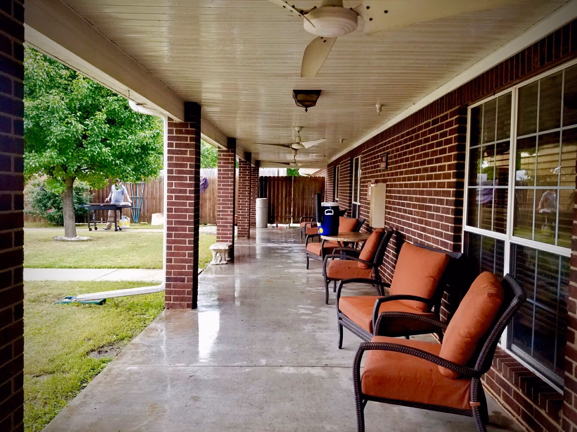 Covered outdoor patio area with brick pillars and walls, featuring several cushioned chairs with orange upholstery lined up along the wall. Ceiling fans are mounted on the white ceiling. In the background, a person is standing near a grill on a grassy lawn with trees and a wooden fence.