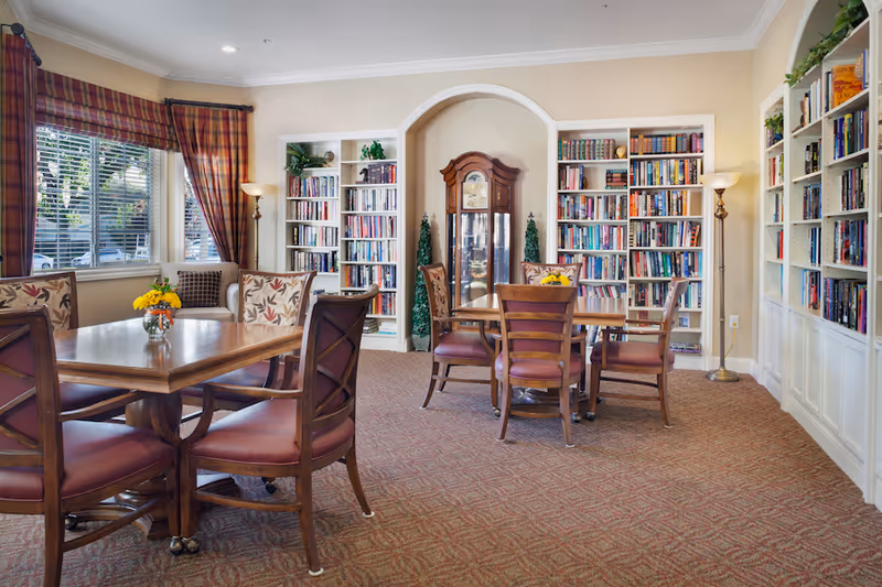 A cozy library room with two wooden tables surrounded by chairs with cushioned seats. The room features built-in white bookshelves filled with books, a grandfather clock in an arched alcove, two floor lamps, and a window with plaid curtains letting in natural light. The carpet is patterned and the walls are painted a light beige color.