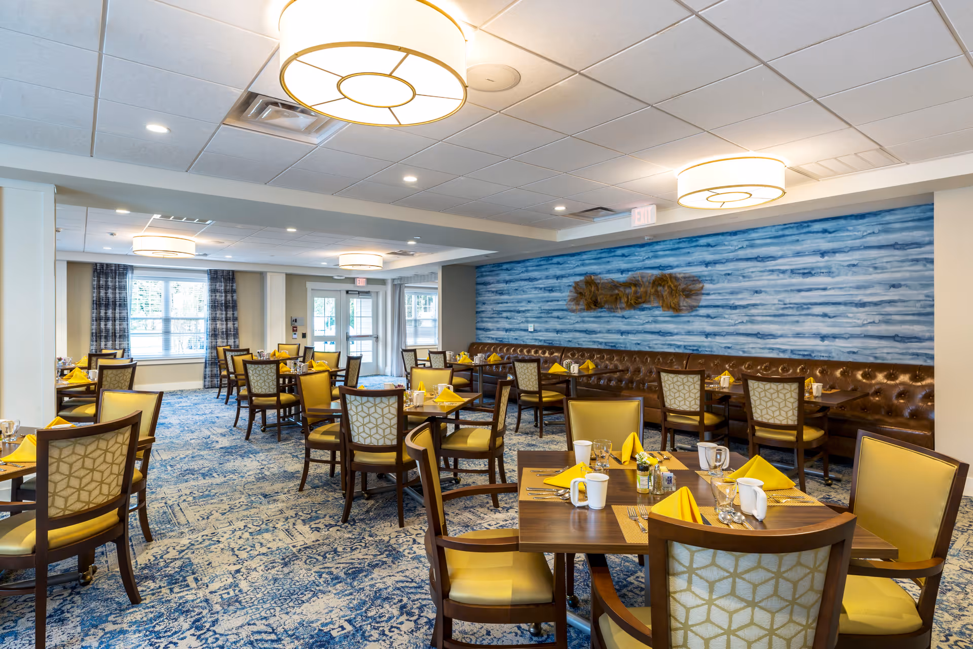 A bright and spacious dining room with multiple wooden tables and chairs. Each table is set with yellow napkins, white mugs, glasses, and silverware. The room features a blue patterned carpet, a blue accent wall with a decorative piece, large windows with plaid curtains, and modern ceiling lights.
