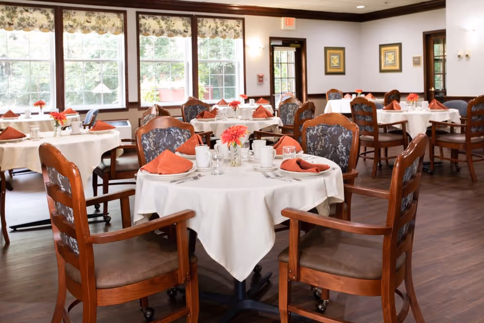 A dining room with multiple round tables covered with white tablecloths, each set with orange napkins, white cups, glasses, and silverware. Wooden chairs with cushioned seats and backs surround the tables. Large windows with floral valances let in natural light, and framed artwork hangs on the walls.