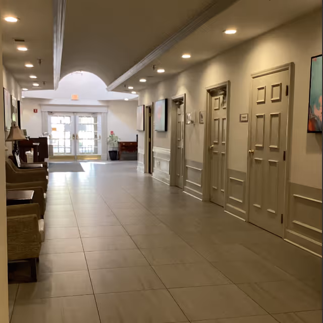 A well-lit hallway in a senior living facility with beige tiled floors, cream-colored walls, multiple closed doors on the right side, and several chairs and small tables along the left wall. The hallway leads to a glass double door entrance with natural light coming through.