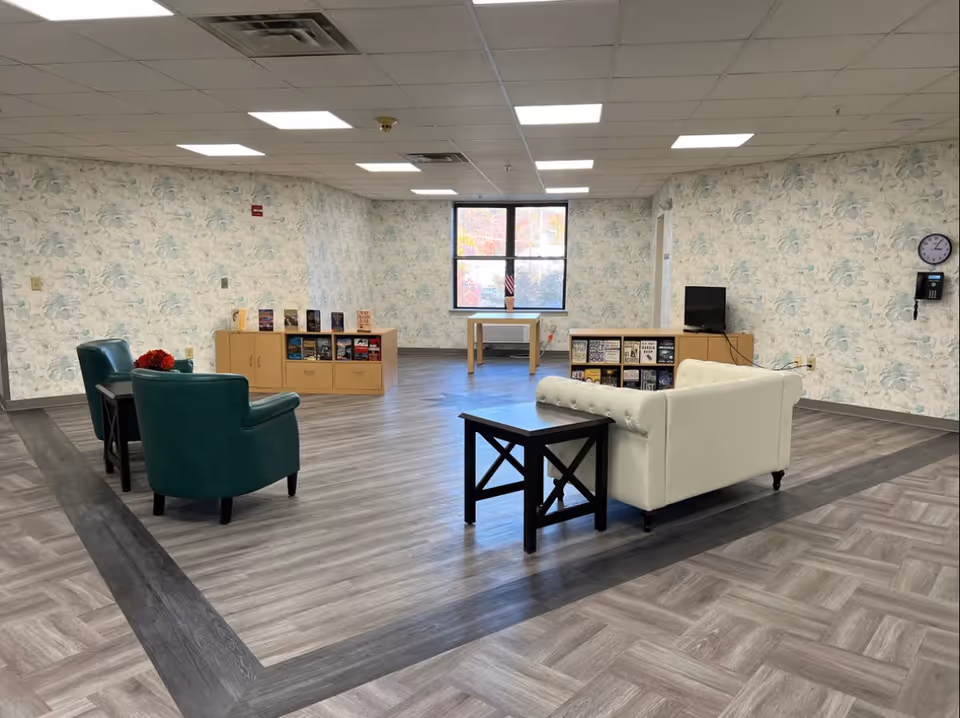 A spacious common area with floral wallpaper, two green armchairs, a white tufted sofa, two black side tables, wooden bookshelves filled with books, a small TV, a wall clock, and a window letting in natural light.