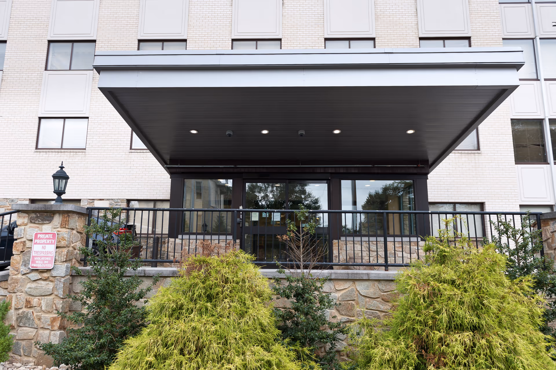 Entrance of a multi-story building with a covered porch supported by stone pillars and black metal railing. There are green shrubs and plants in front of the entrance. The building has light-colored brick walls and several windows. A sign on one stone pillar reads 'Private Property No Trespassing Violators Will Be Prosecuted.'