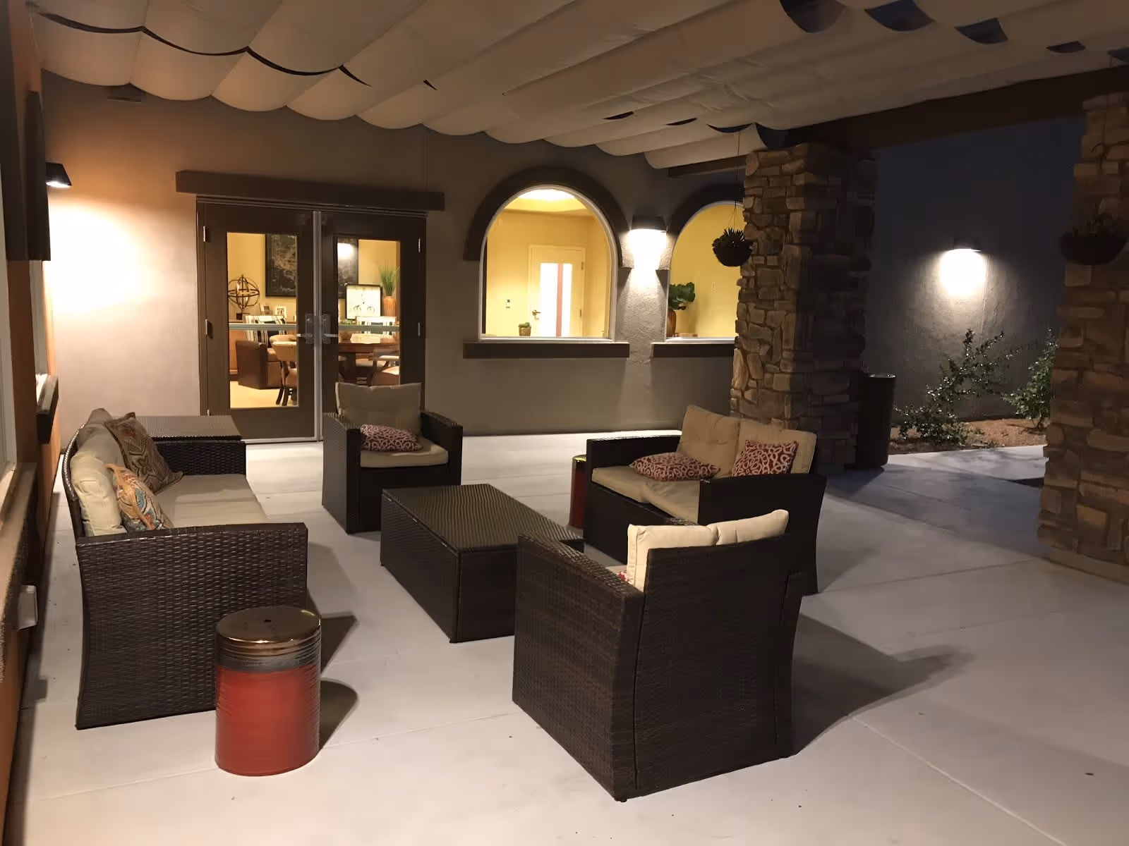 Outdoor covered patio area at night with wicker seating furniture including a sofa, chairs, and a coffee table. The patio has stone pillars and is adjacent to a building with arched windows and glass double doors leading inside to a well-lit room with a dining table and chairs.