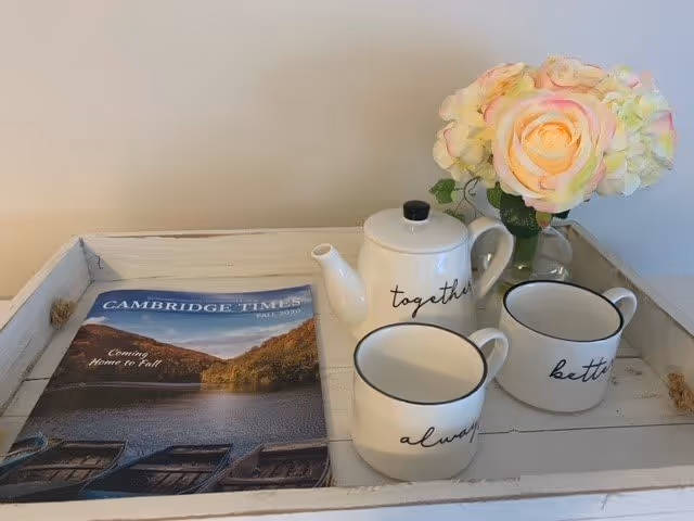 A white wooden tray holding a magazine titled Cambridge Times Fall 2020, a white teapot with the word 'together' written on it, two white mugs with the words 'always' and 'better' written on them, and a small glass vase with a bouquet of light pink and cream-colored flowers.