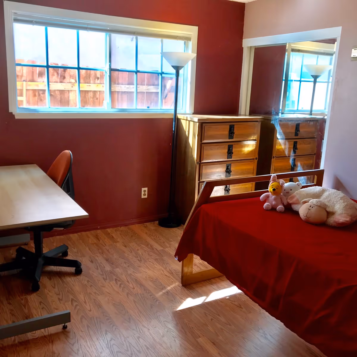 A cozy bedroom with a wooden floor, a bed covered with a red blanket and three stuffed animals on it, a wooden dresser with a mirror behind it, a tall floor lamp, a window with a view of a wooden fence, and a desk with an office chair.