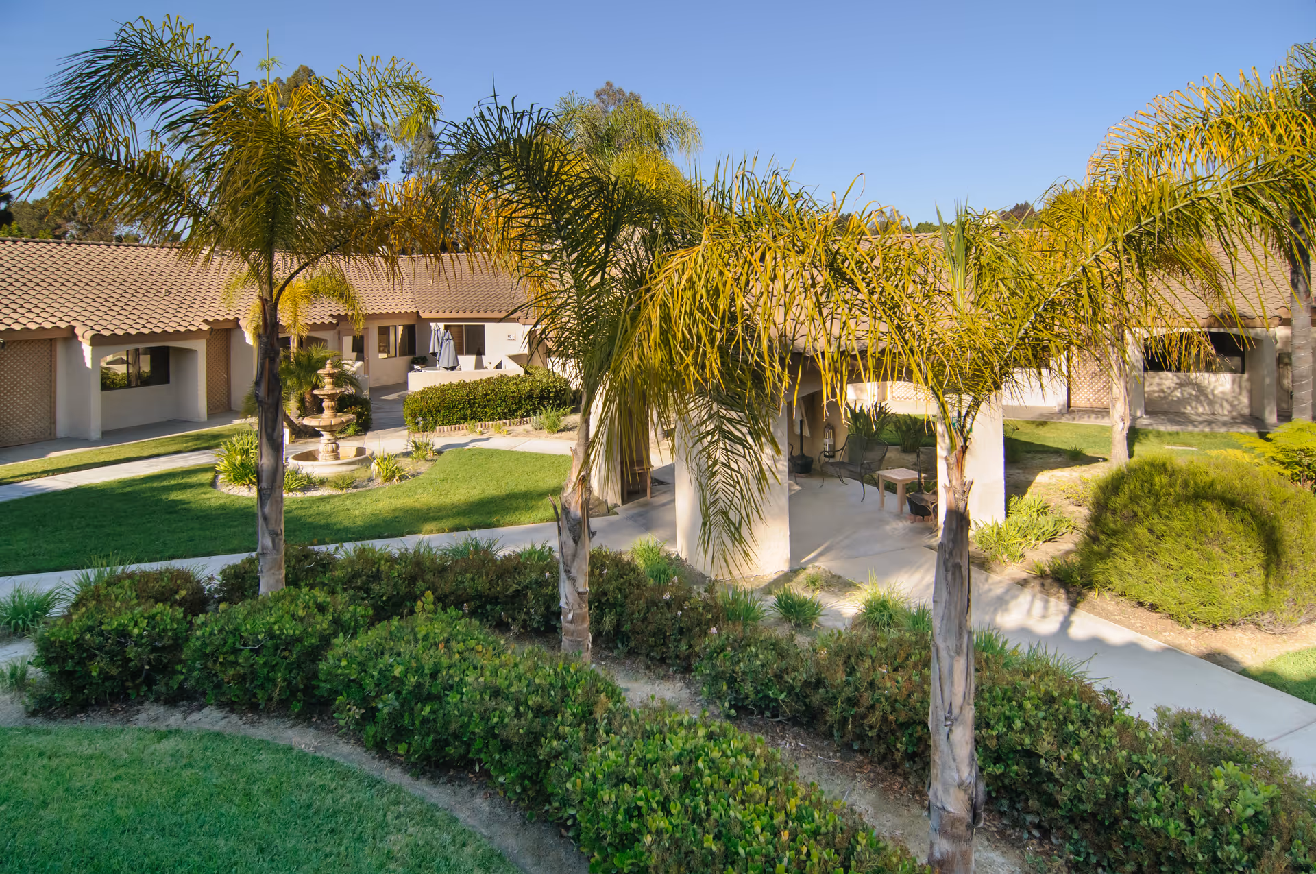 Outdoor courtyard area of a senior living facility with palm trees, green bushes, a fountain, and a covered seating area surrounded by single-story buildings with tiled roofs under a clear blue sky.