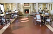 A spacious senior living common area with multiple tables and chairs arranged for dining or socializing. The room features a fireplace in the center with built-in shelves on either side, decorated with various items. The floor is wooden, and the room is well-lit with natural light.