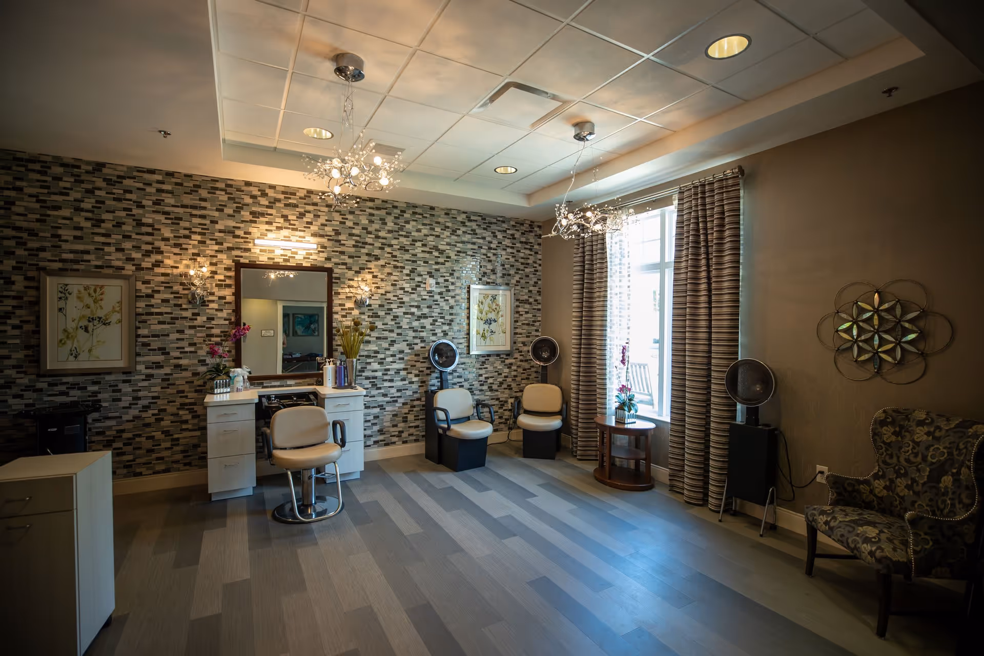A well-lit salon area with a styling chair in front of a mirror and vanity, two hair drying chairs with hooded dryers, a window with striped curtains, decorative wall art, and a patterned armchair.