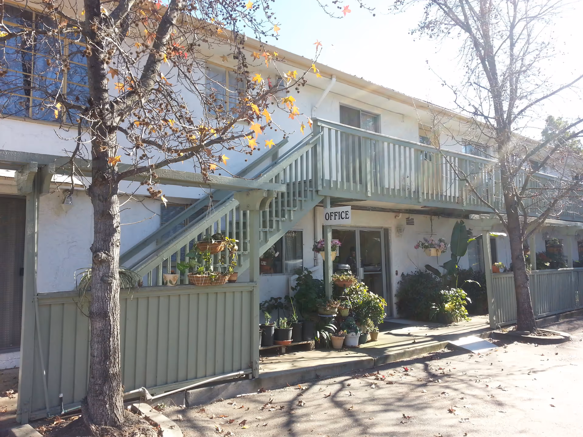 Exterior view of a two-story building with a green wooden staircase and railing leading to the upper floor. There are several potted plants and flowers arranged near the entrance under the staircase. A sign labeled 'OFFICE' hangs above the entrance door. Leafless trees are visible in front of the building, and sunlight casts shadows on the pavement.