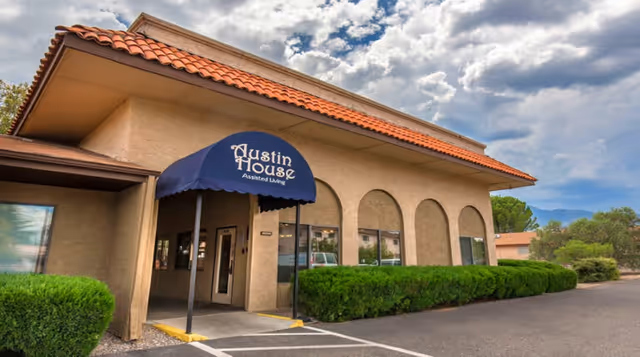 Exterior view of a beige stucco building with a red tile roof and a blue awning that reads 'Austin House Assisted Living' over the entrance. There are trimmed green bushes along the front and a partly cloudy sky overhead.