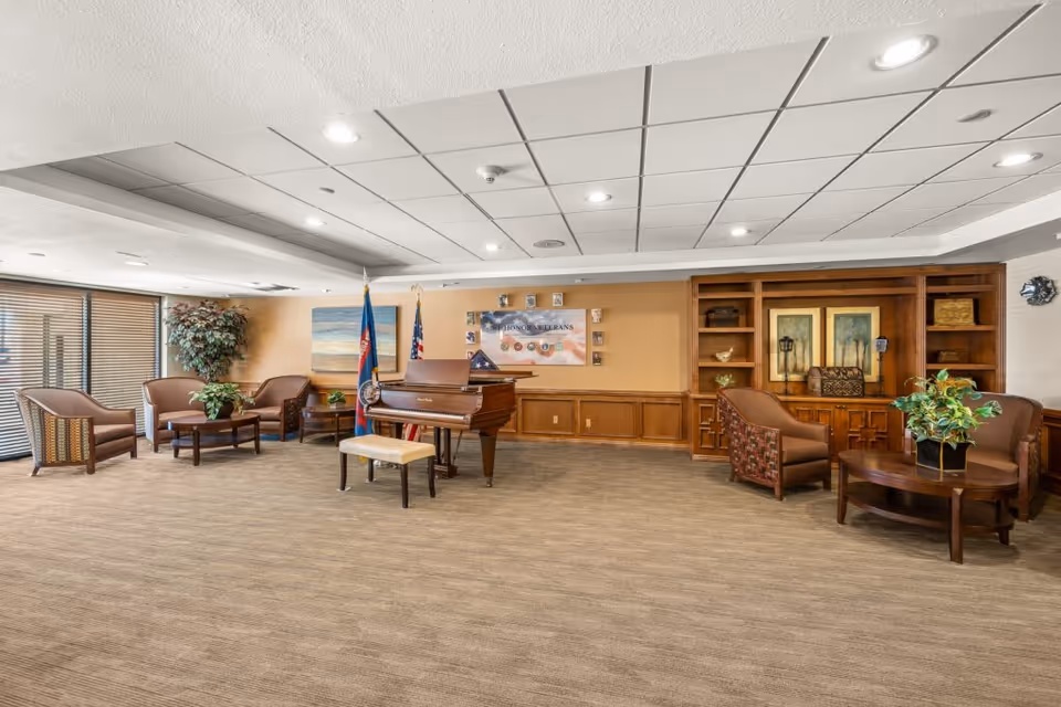 A spacious common area with a grand piano and bench in the center, surrounded by comfortable armchairs and small tables. The room features a carpeted floor, a drop ceiling with recessed lighting, and large windows with blinds on the left side. There are two flags behind the piano and a wall display honoring veterans. Wooden built-in shelves and cabinets with decorative items are on the right side of the room.