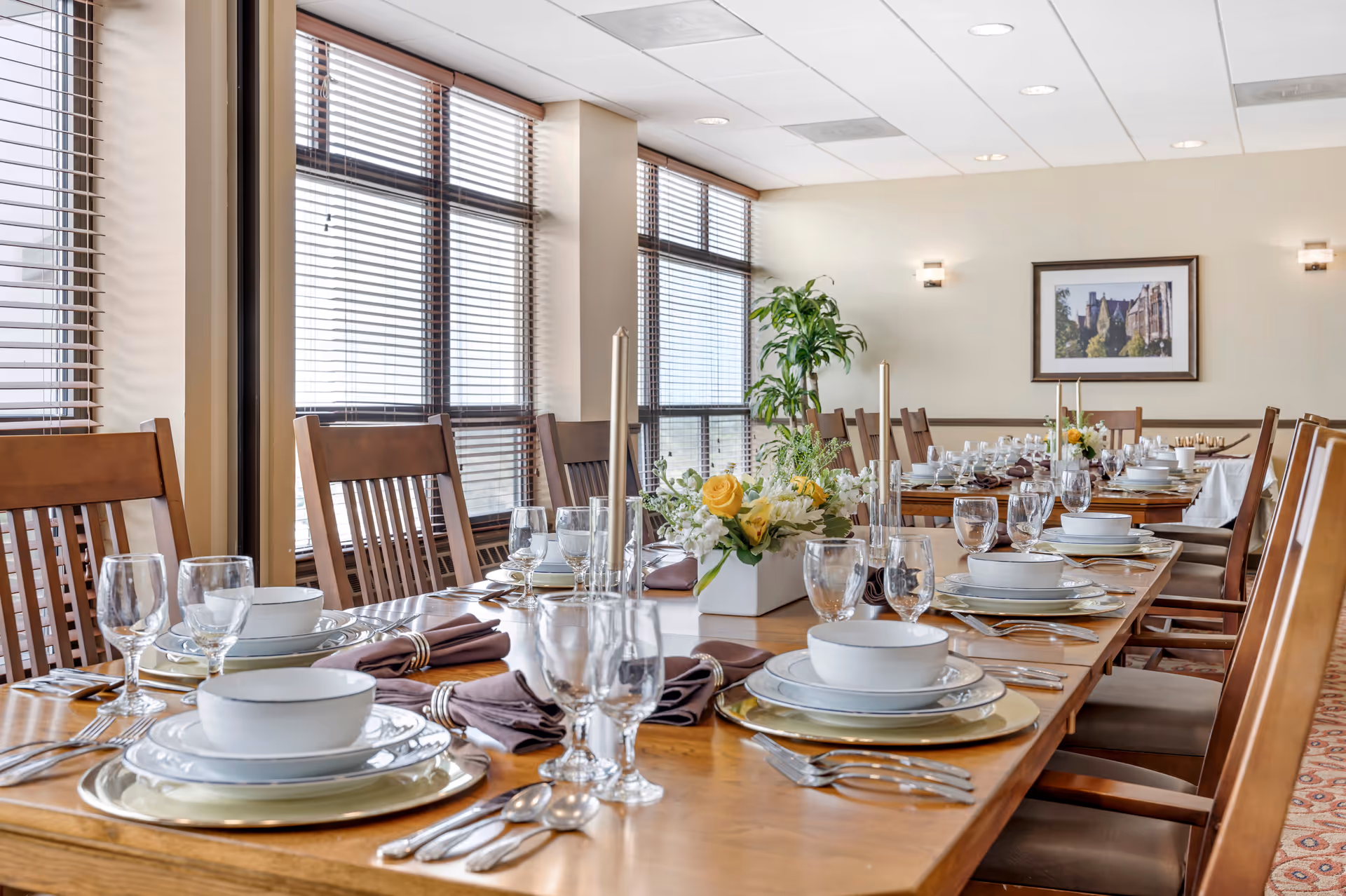 A formal dining room with a long wooden table set with plates, bowls, glassware, napkins and floral centerpieces, surrounded by wooden chairs and large windows.