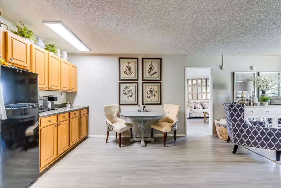 Interior view of a senior living facility kitchen and dining area at Sun City Gardens. The kitchen features wooden cabinets, a black refrigerator, a coffee maker, and a sink. Adjacent to the kitchen is a small round dining table with two upholstered chairs. The room has light-colored flooring and walls decorated with four framed botanical prints. To the right, there is a glimpse of a living room area with a patterned armchair, a white sideboard with a lamp, and decorative plants.