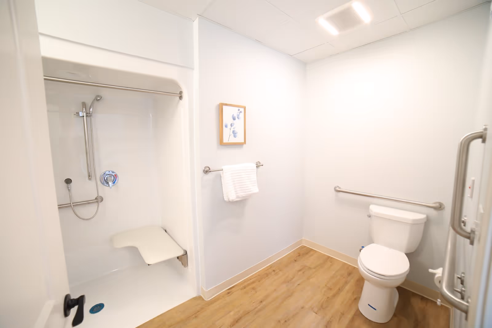 A clean, accessible bathroom with a white toilet, grab bars, a walk-in shower with a fold-down seat, a towel rack with a white towel, light wood flooring, and a small framed artwork on the wall.