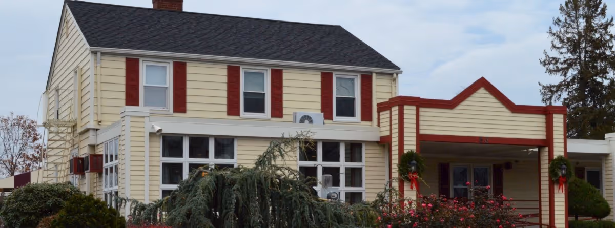 Exterior view of a two-story yellow building with red trim and shutters, surrounded by bushes and trees under a cloudy sky.