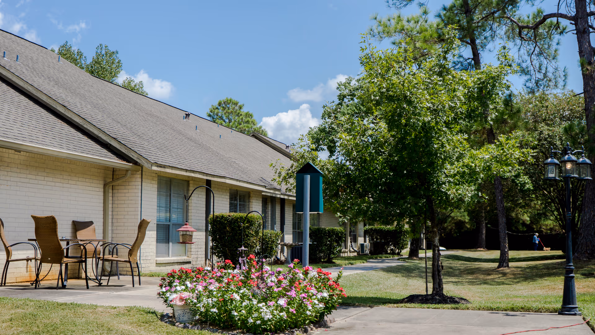 Exterior of a single-story retirement center showing a patio with chairs, a flower bed, walkway and trees on a sunny day.