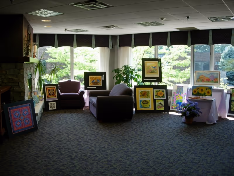 A cozy common area with large windows letting in natural light, featuring two armchairs and a sofa. Several framed paintings and artwork are displayed on the floor and tables near the windows. There is a stone fireplace on the left side and some indoor plants near the windows.