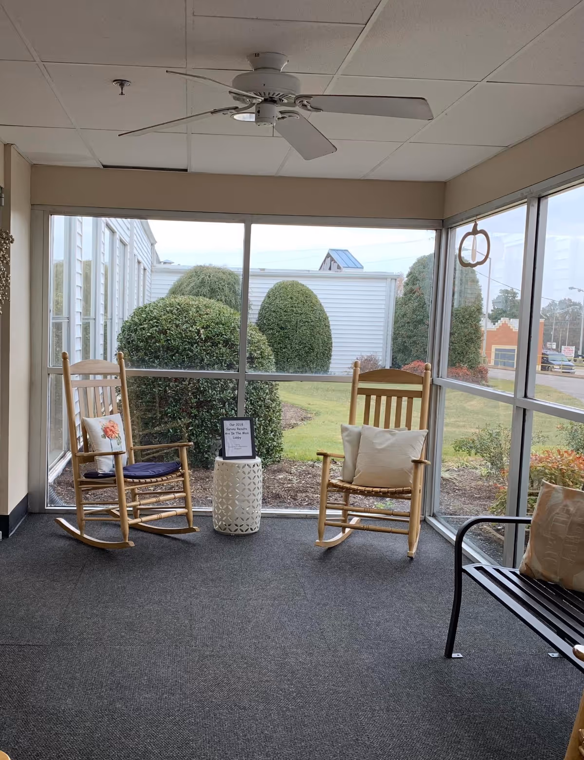 Two wooden rocking chairs and a bench by large windows overlooking trimmed bushes, with a ceiling fan above.