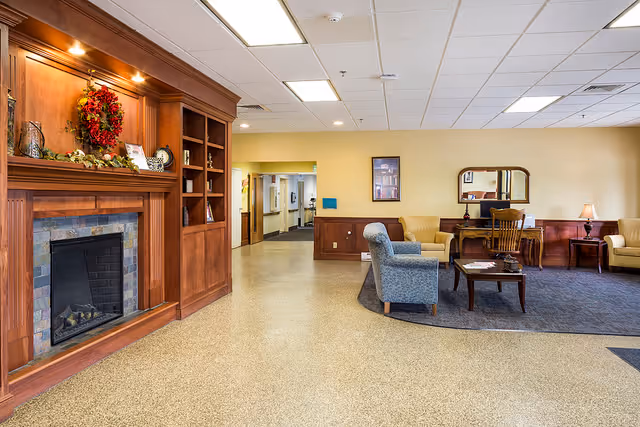 A spacious living room area in a senior living facility featuring a wooden fireplace with a decorative wreath and shelves, comfortable armchairs and sofas arranged around a coffee table on a rug, a wooden desk with a mirror and lamp, and a hallway leading to other rooms.